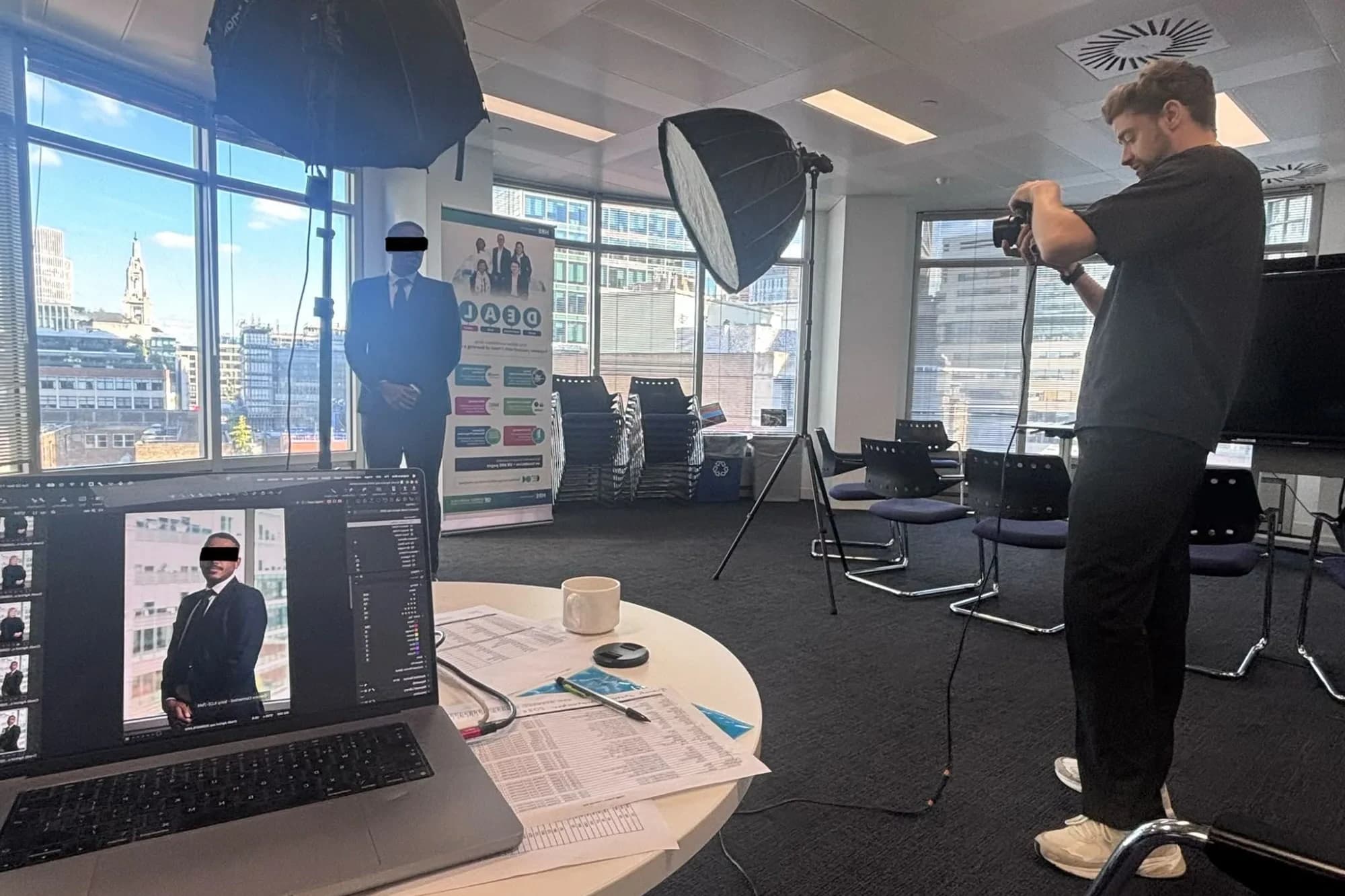 Photographer Etienne Morax shooting a staff headshot in Crédit Agricole CIB's London office, with two softboxes, a City skyline backdrop and a tethered laptop showing the portrait